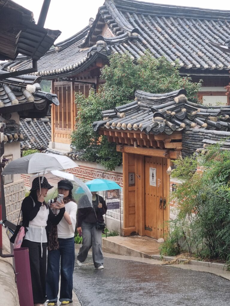 Scenic close-up of traditional Korean hanok tiled roofs and patterned walls at Bukchon Hanok Village with visitors walking under umbrellas in the rain